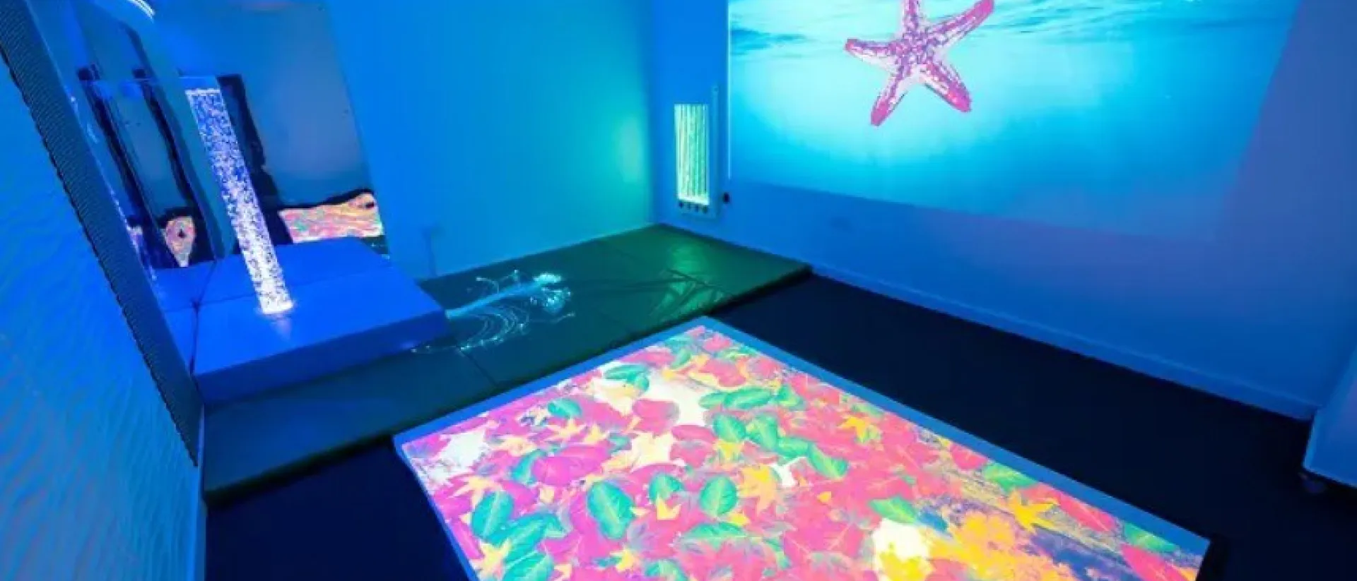 An image showing a sensory room with a bubble tube, starfish projected on screen and a light up floor